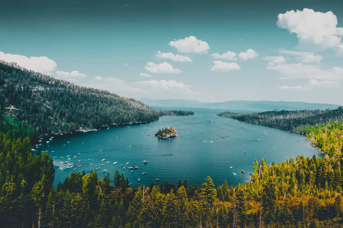 Aerial view of a forested lake with boats and a small island, surrounded by dense pine trees and a clear sky.